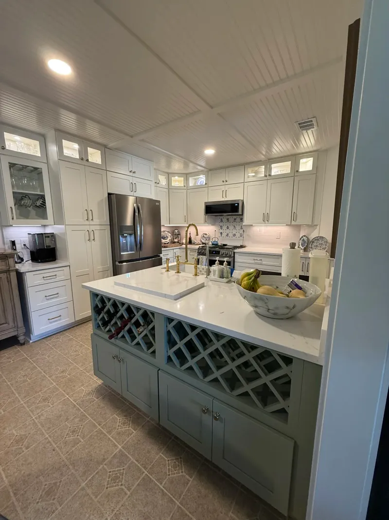 Renovated kitchen with sage green island featuring built-in wine storage, white shaker perimeter cabinets, brushed brass fixtures, and beadboard ceiling.
